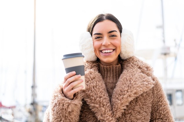 Giovane donna che indossa manicotti invernali e tiene il caffè da asporto all'aperto sorridendo molto