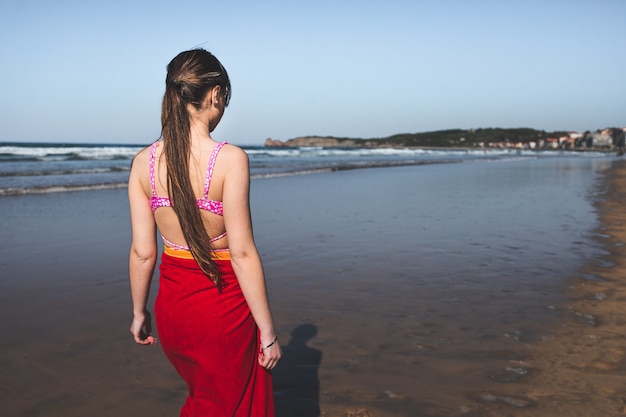 Giovane donna che indossa un asciugamano rosso e un bikini rosa che cammina al bordo dell'acqua sulla spiaggia