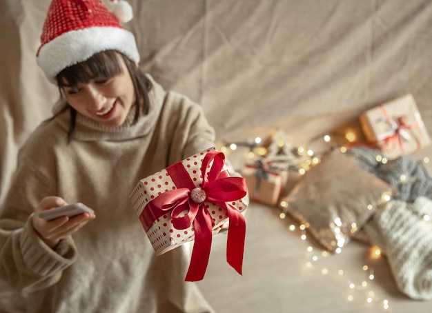 Giovane donna che indossa un cappello di Babbo Natale in possesso di un regalo di Natale splendidamente avvolto. Concetto di celebrare il Natale durante la pandemia di coronavirus.