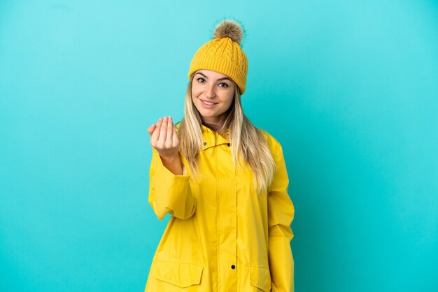 Giovane donna che indossa un cappotto antipioggia su sfondo blu isolato che invita a venire con la mano. Felice che tu sia venuto