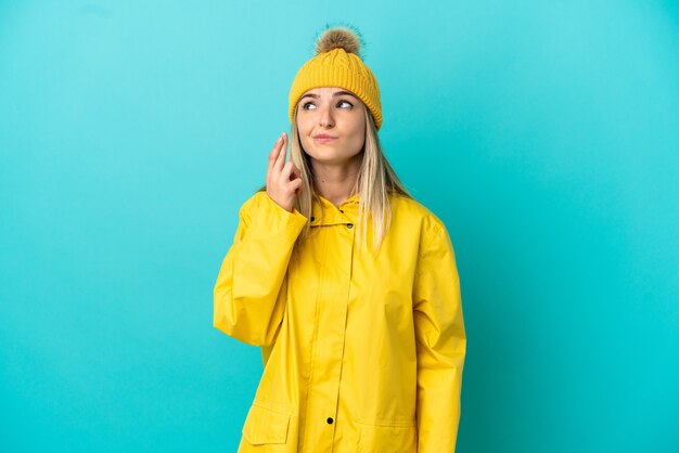 Giovane donna che indossa un cappotto antipioggia su sfondo blu isolato con le dita incrociate e augurando il meglio