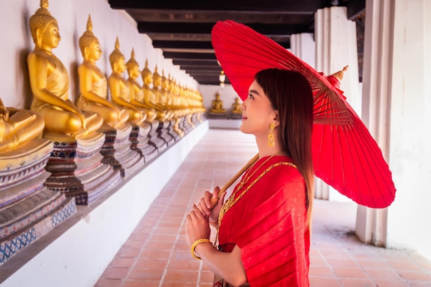 Giovane donna che indossa un tradizionale abito thailandese rosso e accessori dorati tiene in mano un ombrello tradizionale nel sito storico Wat Phutthaisawan Ayutthaya costume nazionale thailandese