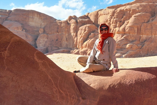 Giovane donna che indossa una giacca calda e una sciarpa intorno alla testa sorridente, seduta alla finestra di roccia formata naturalmente nel deserto di wadi rum in una giornata di sole