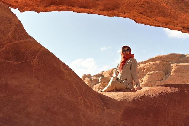 Giovane donna che indossa una giacca calda e una sciarpa intorno alla testa sorridente, seduta alla finestra di roccia formata naturalmente nel deserto di Wadi Rum (Valle della Luna) in una giornata di sole
