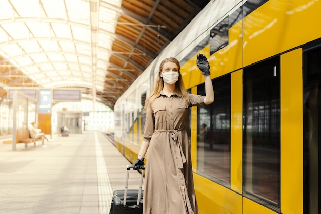 Giovane donna che indossa una maschera facciale e guanti e saluta in una stazione ferroviaria - COVID-19