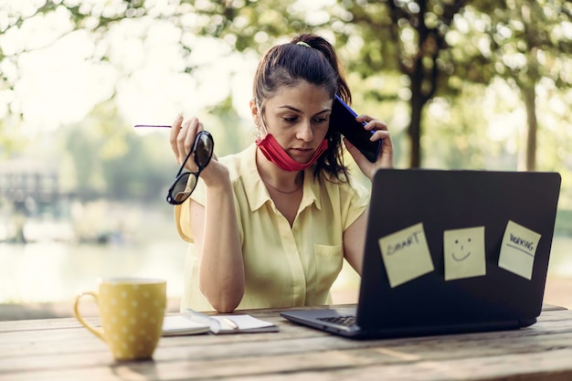 Giovane donna che indossa una maschera facciale protettiva abbassata utilizzando computer portatile e smartphone all'aperto Libero professionista femminile che lavora su laptop al parco Lavoro a distanza nuovo concetto normale durante il tempo covid19