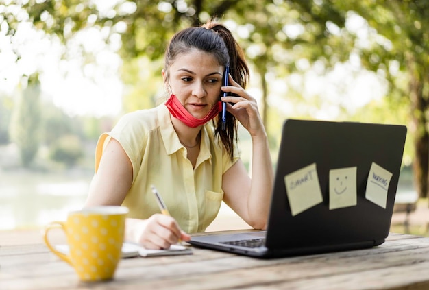 Giovane donna che indossa una maschera facciale protettiva abbassata utilizzando un computer portatile all'aperto Libero professionista o manager femminile che lavora su un computer portatile al parco Lavoro a distanza e nuovo concetto normale durante il tempo del coronavirus