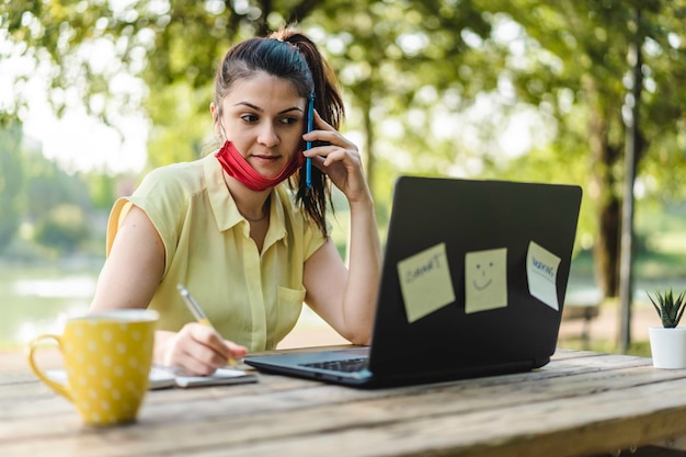 Giovane donna che indossa una maschera protettiva aperta utilizzando il laptop all'aperto Imprenditrice femminile che prende appunti mentre chiama con il cellulare al parco Lavoro a distanza nuovo concetto normale dopo la pandemia di covid 19