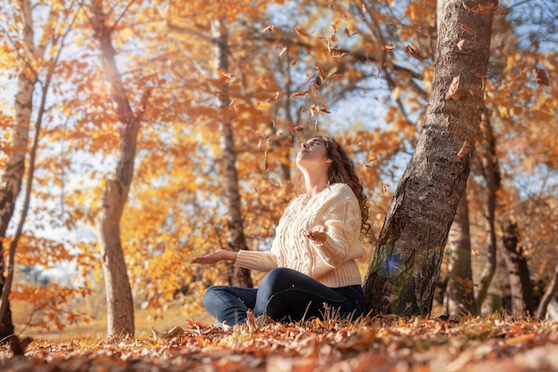 Giovane donna che lancia foglie gialle nell'aria seduta nella foresta autunnale
