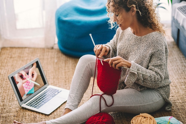 Giovane donna che lavora a maglia la lana usando l'ago mentre guarda un tutorial online sul portatile a casa. Donna che guarda lezioni di cucito, laptop e hobby domestici. Donna caucasica che impara a lavorare a maglia online