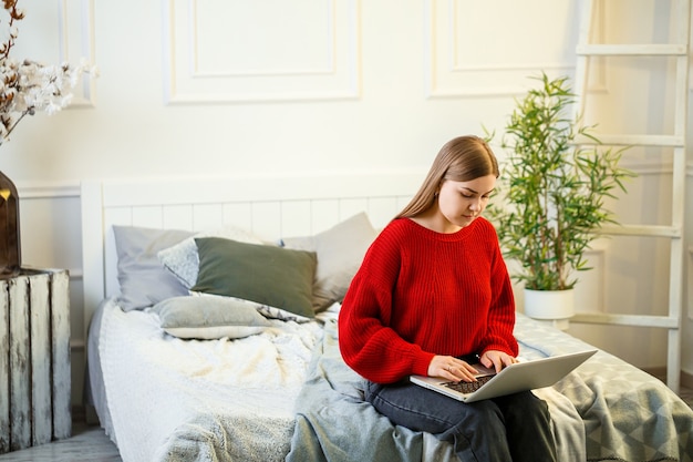 Giovane donna che lavora al computer portatile che si siede sul letto a casa. Lavorare da casa durante la quarantena. Ragazza in maglione e jeans a casa sul letto