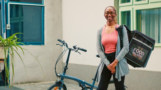 Giovane donna che lavora come trasportatore con la bicicletta, utilizzando lo zaino per consegnare il pacchetto del pasto in un sacchetto di carta. Corriere da asporto femminile in bicicletta per dare ordini di fast food alla porta d'ingresso. Tiro a mano libera.