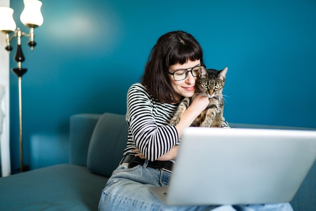Giovane donna che lavora con il computer portatile e con un gatto seduto nel soggiorno moderno a casa