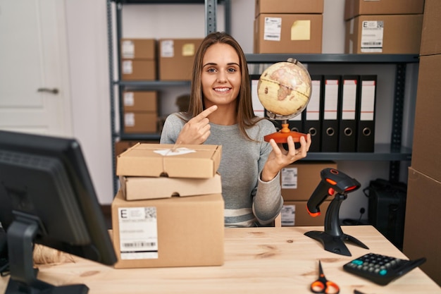 Giovane donna che lavora nel commercio elettronico di piccole imprese tenendo la palla del mondo sorridente felice che punta con la mano e il dito