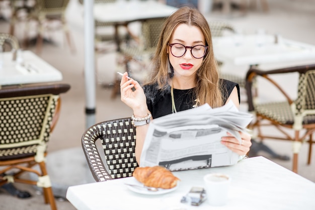 Giovane donna che legge il giornale mentre è seduto con la sigaretta durante la colazione all'aperto nella tipica terrazza del caffè francese in Francia