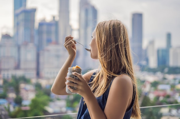 Giovane donna che mangia budino di chia sul suo balcone con vista sulla grande città