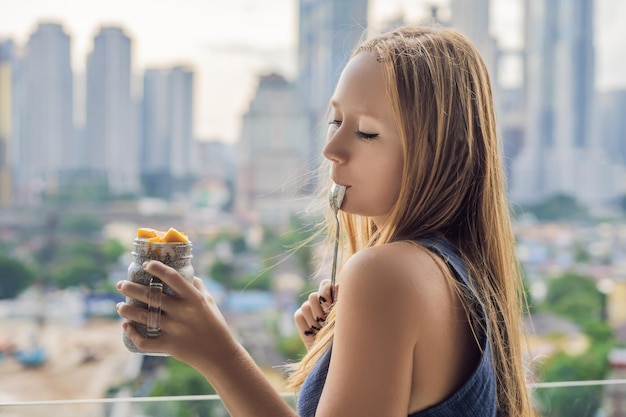 Giovane donna che mangia budino di chia sul suo balcone con vista sulla grande città