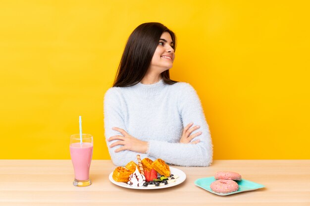 Giovane donna che mangia i waffles e il frappé in una tavola sopra la parete gialla isolata che guarda al lato