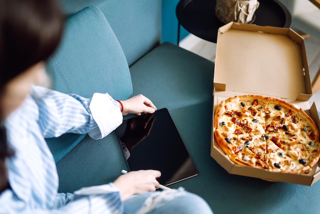 Giovane donna che mangia un pezzo di pizza deliziosa dalla scatola, seduta sul divano di casa. Consegna del cibo.