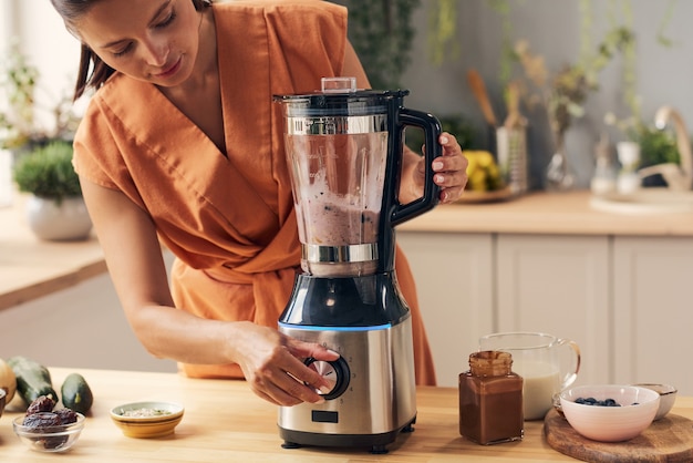 Giovane donna che mescola gli ingredienti nel frullatore elettrico mentre prepara il frullato