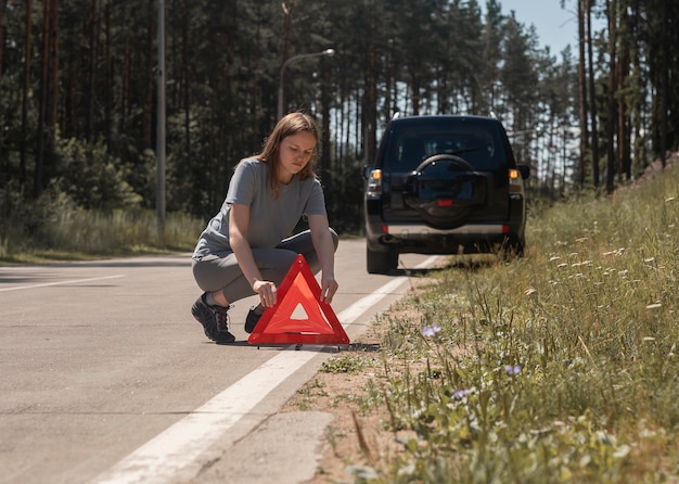 Giovane donna che mette il segno di attenzione del triangolo rosso sulla strada vicino all'automobile rotta sul bordo della strada