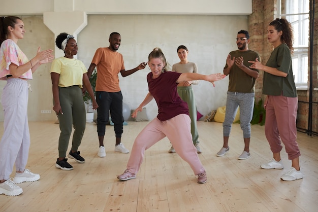 Giovane donna che mostra esercizi di danza mentre altre persone in piedi in cerchio battendo le mani e guardando in studio di danza