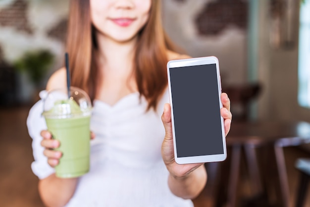 Giovane donna che mostra uno smartphone a schermo vuoto con copia spazio mentre si gusta un drink in una caffetteria