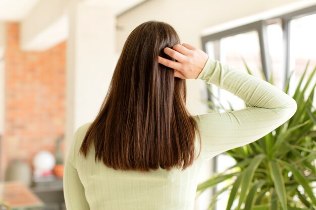 Giovane donna che pensa o dubita di graffiare la testa sentendosi perplessa e confusa indietro o vista posteriore