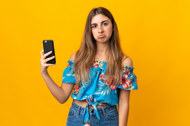 Giovane donna che per mezzo del telefono cellulare sopra fondo giallo isolato con l'espressione triste