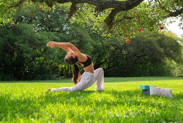 Giovane donna che pratica yoga facendo falce di luna pongono sotto un albero in un concetto di ParkWellness