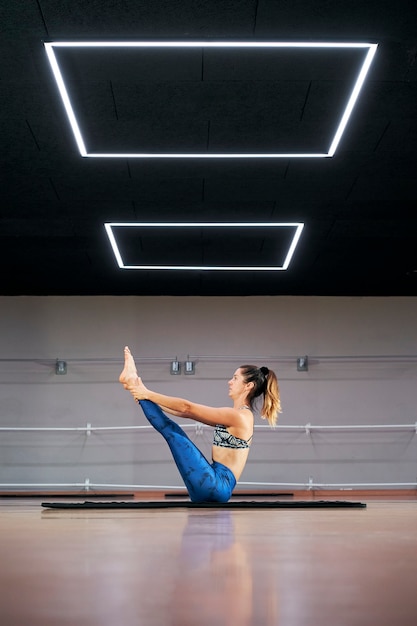 Giovane donna che pratica yoga o pilates in una palestra, esercita in abbigliamento sportivo blu, facendo navasana o posa in barca