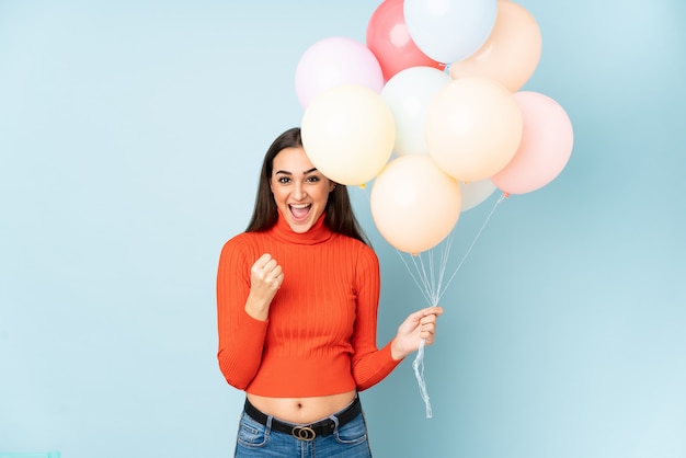 Giovane donna che prende molti palloni isolati sulla parete blu che celebra una vittoria