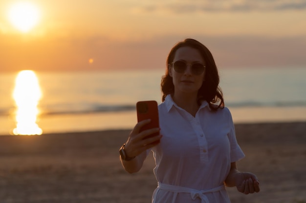 Giovane donna che prende selfie tramite smartphone mentre si trova in spiaggia durante il tramonto