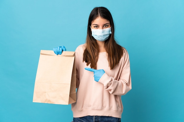 Giovane donna che protegge dal coronavirus con una maschera e in possesso di un sacchetto della spesa isolato su sfondo blu e indicandolo