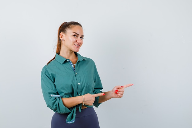 Giovane donna che punta a destra in camicia verde e sembra allegra. vista frontale.