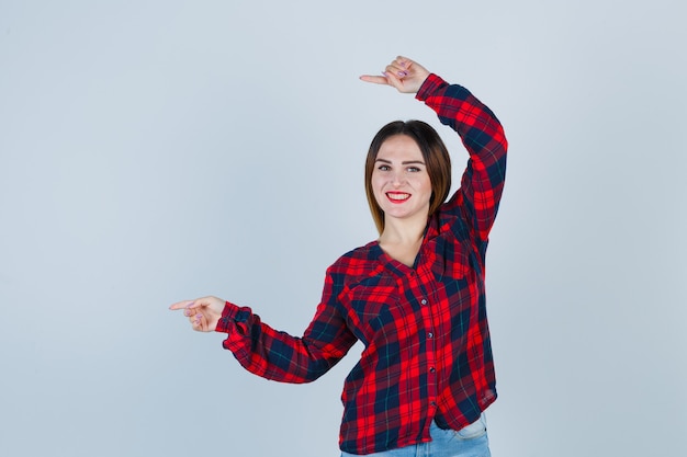 Giovane donna che punta a sinistra con l'indice, sorridente in camicia a quadri, jeans e sembra allegra, vista frontale.