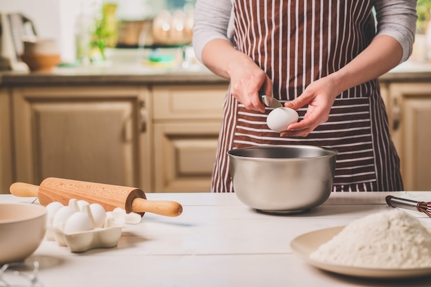 Giovane donna che rompe l'uovo sopra la ciotola con pasta, primo piano. Una donna con un grembiule a righe sta cucinando in cucina