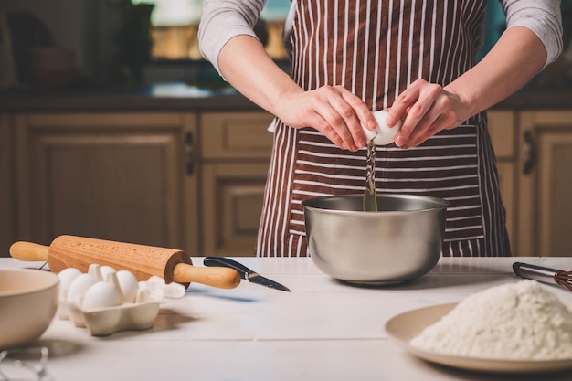 Giovane donna che rompe l'uovo sopra la ciotola con pasta, primo piano. Una donna con un grembiule a righe sta cucinando in cucina