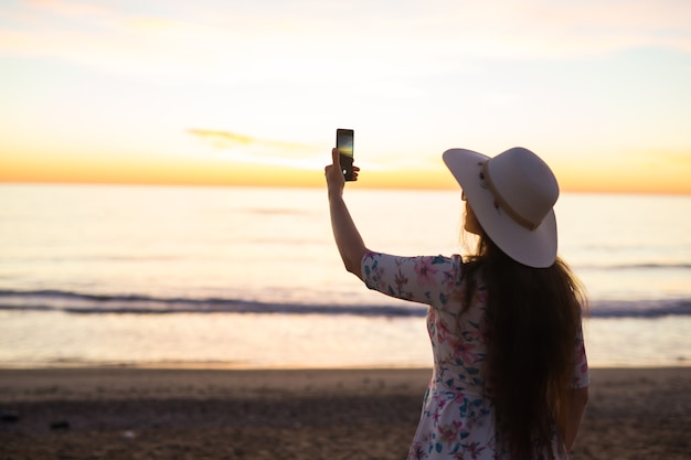 Giovane donna che scatta foto al mare e al tramonto con un telefono cellulare o una fotocamera digitale per smartphone per posta a