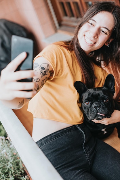 Giovane donna che scatta foto selfie coccole e si prende cura del suo bulldog francese con la lingua fuori casaCucciolo osservando concentrato guardando la cittàGiocoso partner di amicizia umoristico e allegro