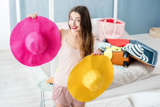 Giovane donna che sceglie cappello in camera da letto con valigia e vestiti sullo sfondo. Preparativi per le vacanze estive