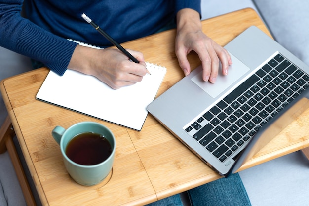 Giovane donna che scrive a matita allo scrittorio di legno con il computer portatile e la tazza di tè.