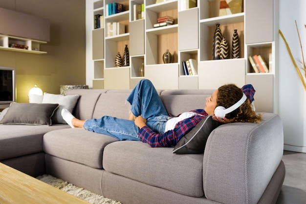 Giovane donna che si distende nel soggiorno o nella tana a casa su un divano ascoltando musica con le cuffie stereo