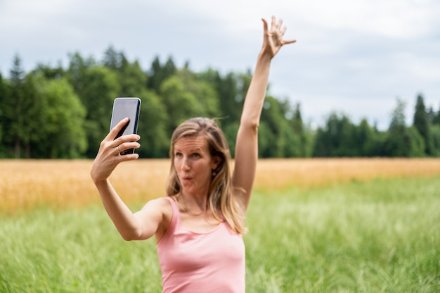 Giovane donna che si gode la natura facendo selfie mentre solleva il braccio e fa una faccia buffa. Con focus sul telefono cellulare.