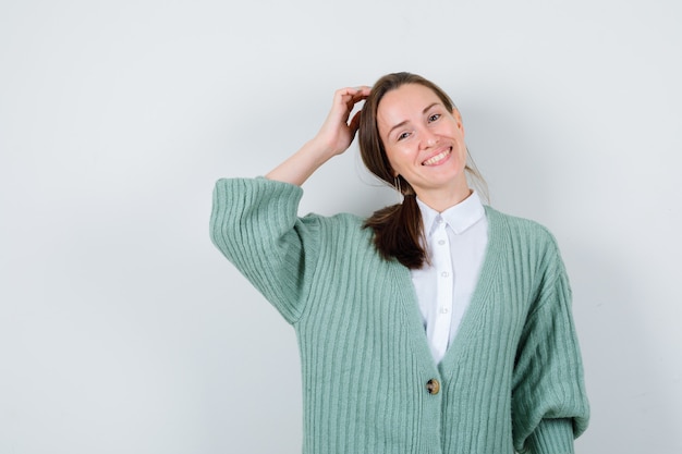 Giovane donna che si gratta la testa in camicetta, cardigan e sembra allegra, vista frontale.