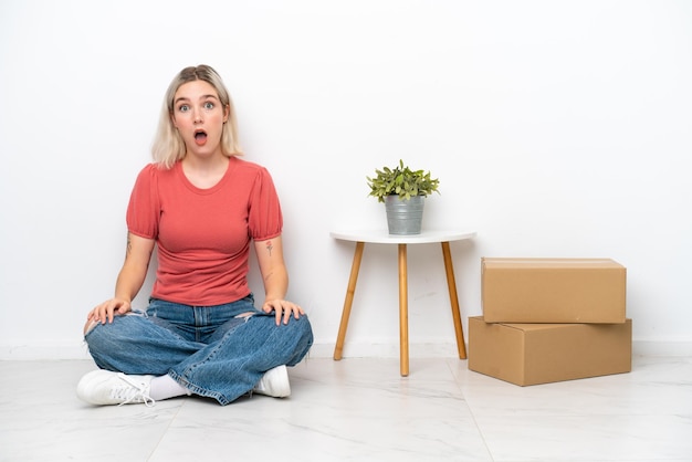 Giovane donna che si muove in una nuova casa tra scatole isolate su sfondo bianco con espressione facciale a sorpresa