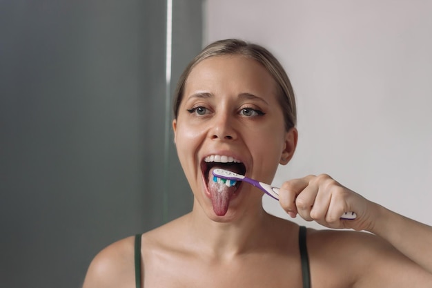 Giovane donna che si pulisce la lingua dalla placca bianca usando uno spazzolino da denti guardandosi nello specchio del bagno