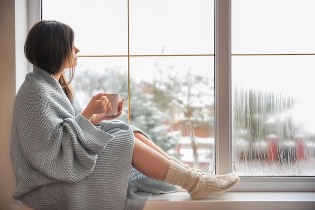 Giovane donna che si rilassa bevendo il tè vicino alla finestra il giorno d'inverno