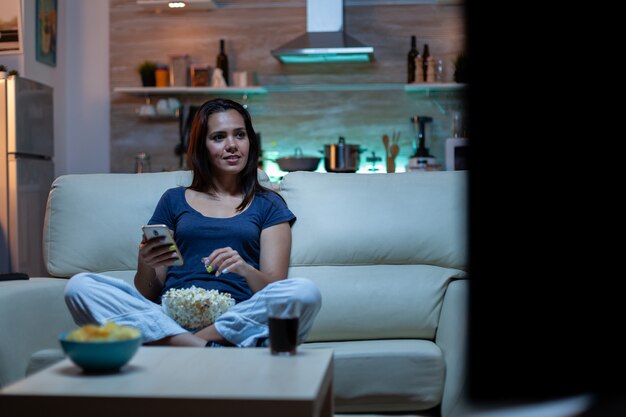 Giovane donna che si rilassa di notte usando il telefono e guardando la tv seduta sul divano. Donna felice divertita sola che legge, scrive, cerca, naviga su smartphone ridendo divertendosi usando la tecnologia internet