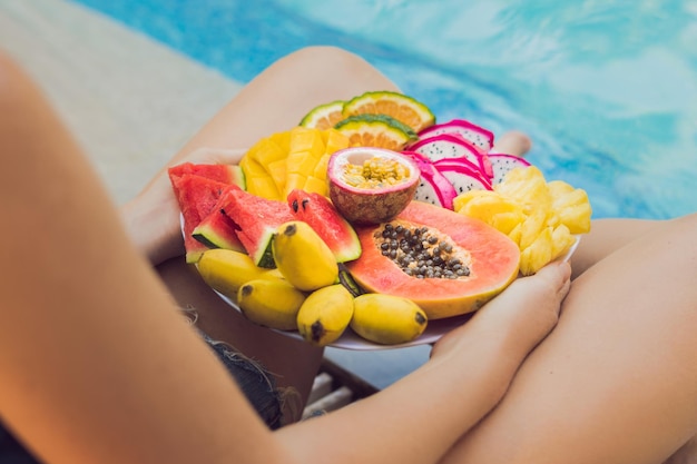 Giovane donna che si rilassa e mangia un piatto di frutta a bordo piscina dell'hotel. Dieta estiva esotica. Foto di gambe con cibo sano a bordo piscina, vista dall'alto. Stile di vita da spiaggia tropicale.